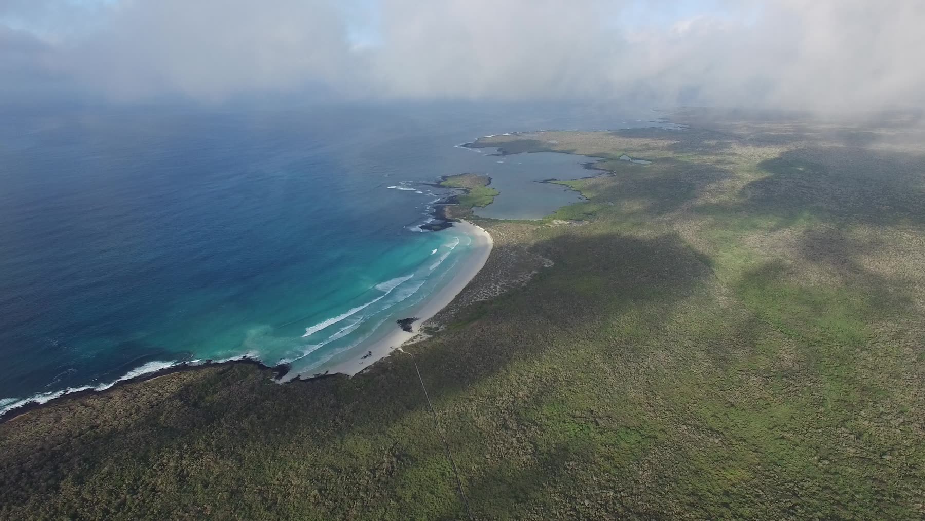 Empty white-sand cove with turquoise water, backed by dark lava rock and scrub